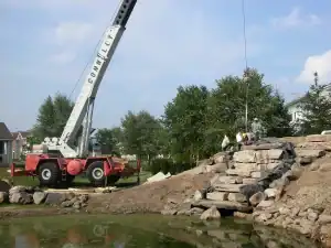 Crane installing large stones for outdoor landscape and stone wall building.